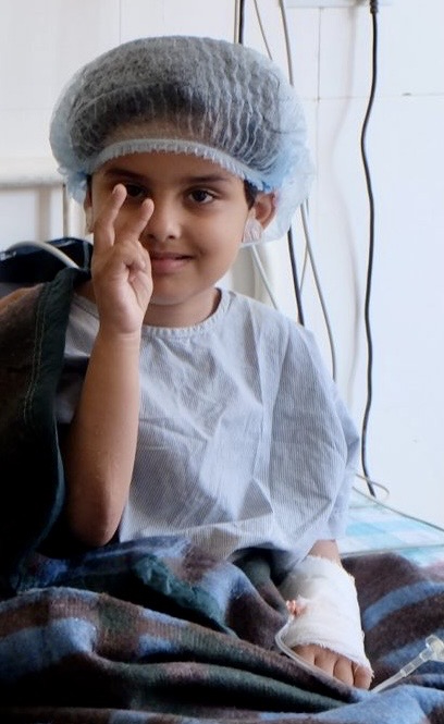 Ein Junge mit OP-Haube und verbundenem Arm sitzt im Krankenhausbett und hebt zwei Finger.